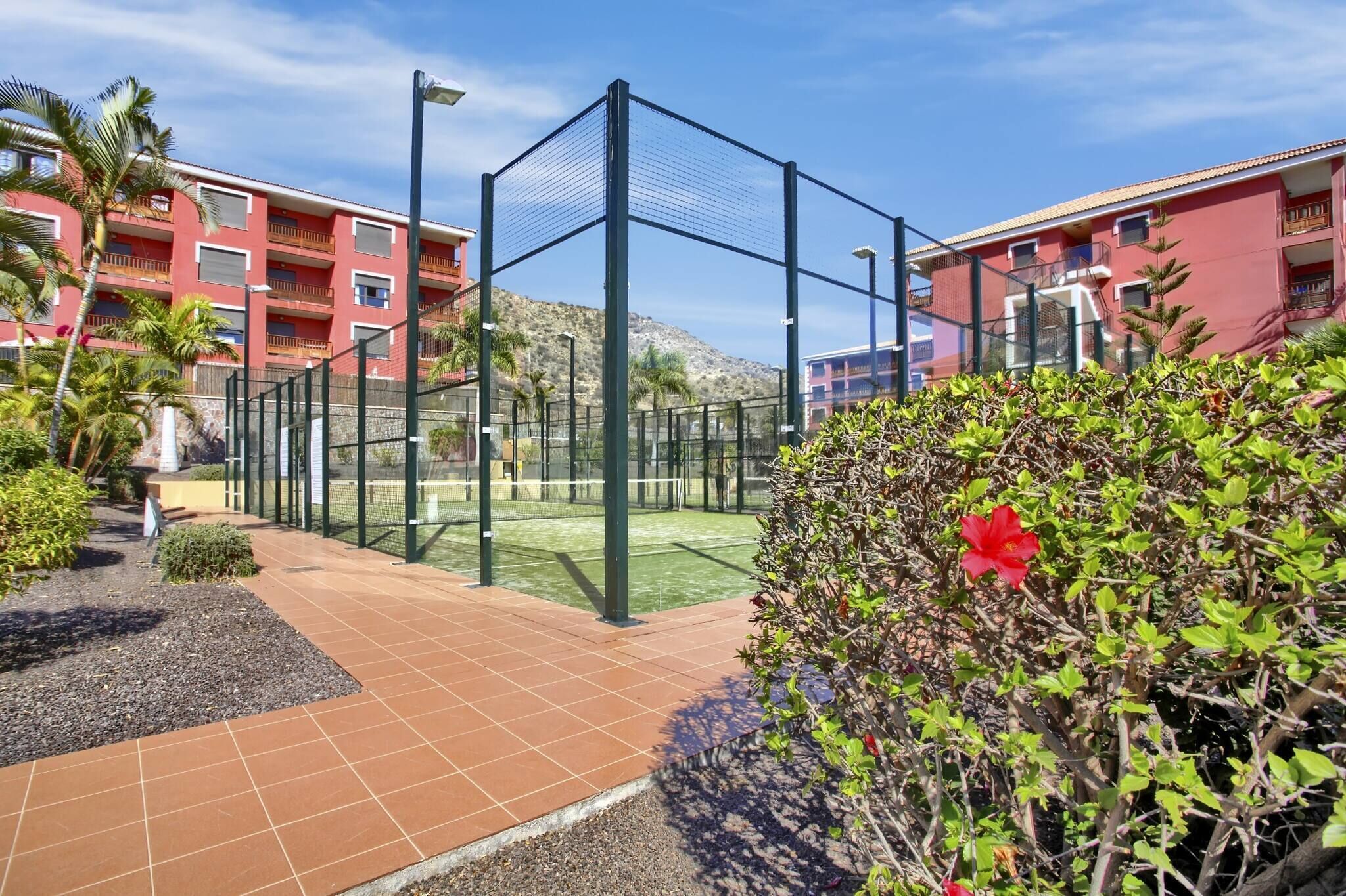A tennis court surrounded by a fence, with a red building and palm trees in the background.