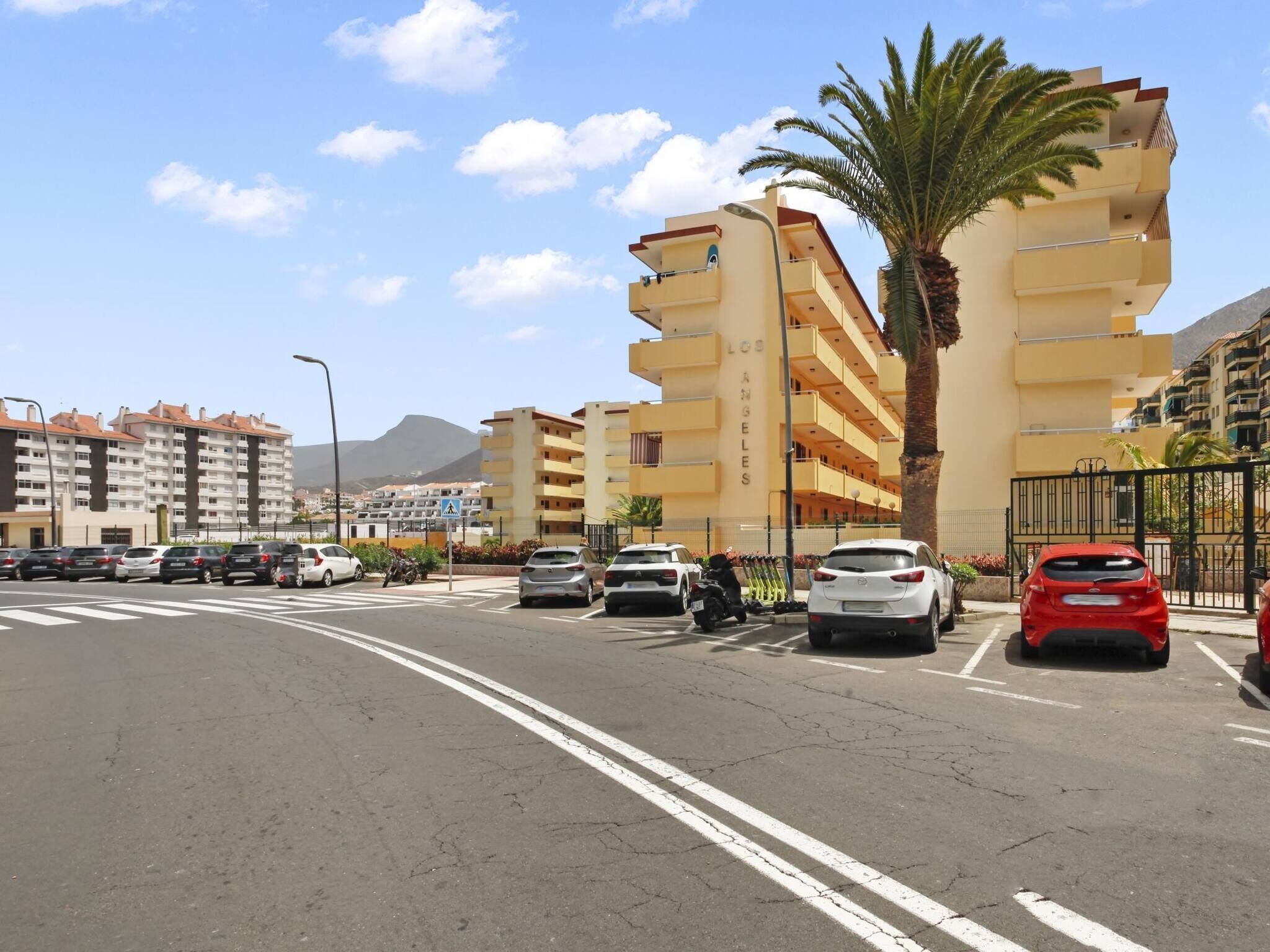 A multi-story residential building with balconies, a palm tree, and parked cars on a sunny day.