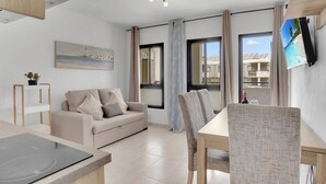 A modern living room with a sofa, dining table, and chairs. There is a TV mounted on the wall and a window with a view of buildings. - Ferdy's Home, Pool, Wi Fi, Downtown Los Cristianos (Los Cristianos)