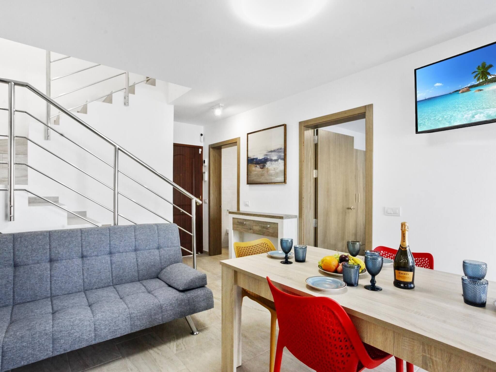 Ein modernes Wohnzimmer mit einem grauen Sofa, einem Holzesstisch und roten Stühlen. Eine Treppe führt zu einer weiteren Ebene.