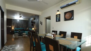 A dining area with a glass table and wooden chairs, a ceiling fan, and an air conditioning unit. - Banipark Service Apartment (Jaipur)
