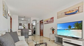 Ein modernes Wohnzimmer mit Flachbildfernseher, einer Couch, einem Essbereich und einer Küche im Hintergrund.