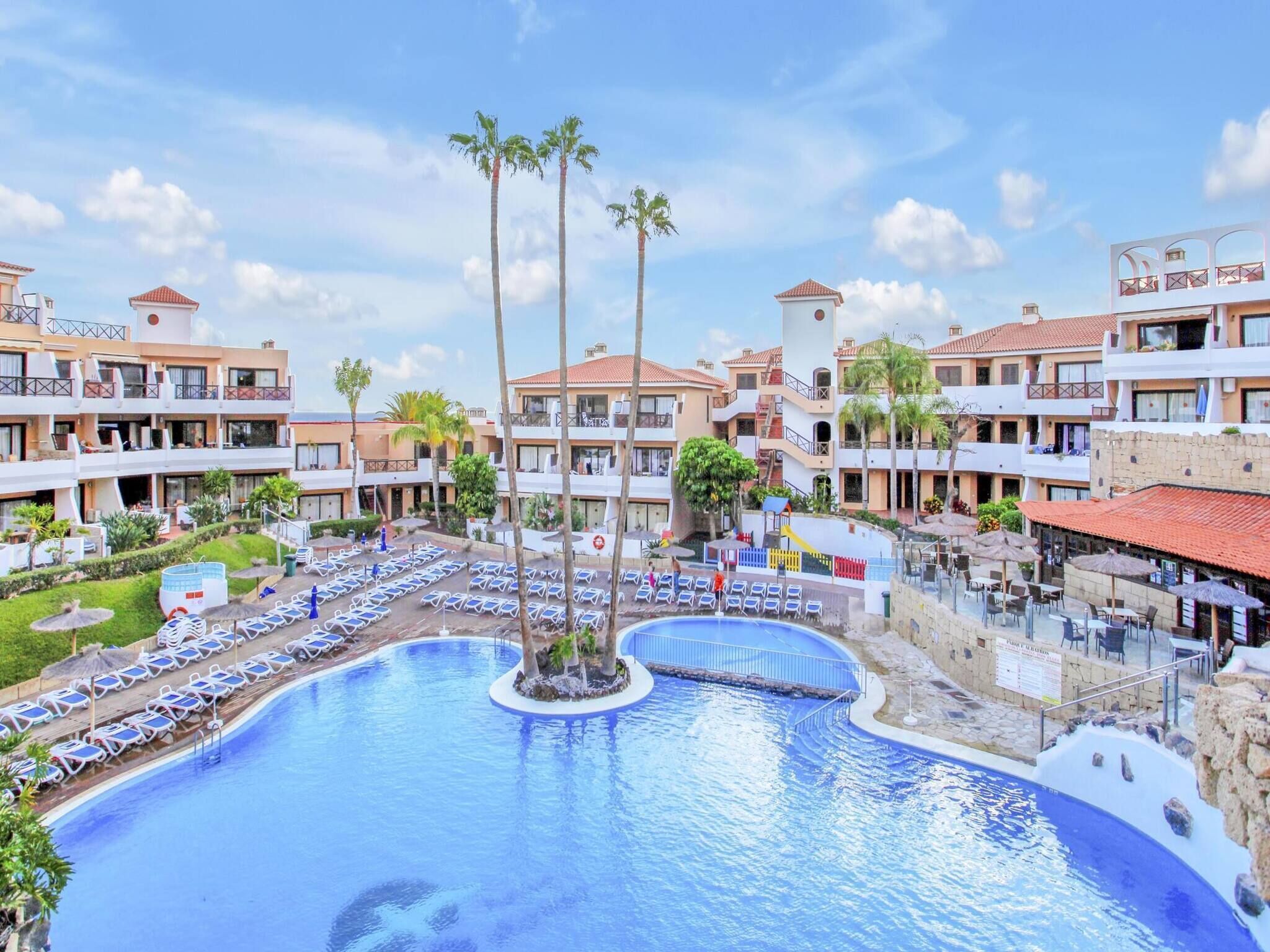 A resort with a central swimming pool surrounded by lounge chairs and palm trees.