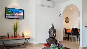 A modern living room with a flat-screen TV, a Buddha statue, and a dining area with a table and chairs.