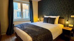 Een net opgemaakt bed met een donkere hoofdbord, een raam met gordijnen, en een behang met patroon.