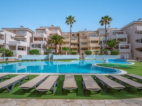 Une piscine entourée de chaises longues, de palmiers et d’un immeuble à plusieurs étages doté de balcons.