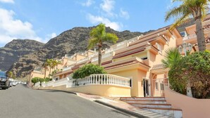 A coastal street with a row of buildings, a clear sky, and mountains in the background. - The Stone Apartment, Los Gigantes, Heated Pool (Acantilados de Los Gigantes)