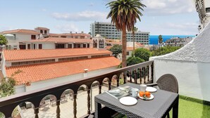 A balcony with a table set for two, overlooking a cityscape with terracotta-roofed buildings and a palm tree.