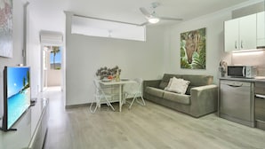 Una sala moderna con una televisión de pantalla plana, una mesa de comedor con sillas, un sofá, un ventilador de techo y una zona de cocina con microondas y lavavajillas.