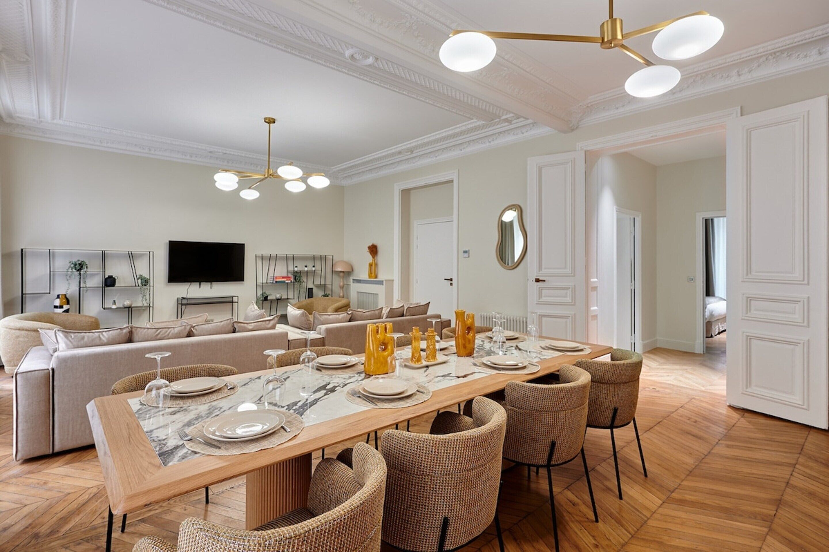 A spacious living room with a long dining table set for six, a sofa, and a television in the background.