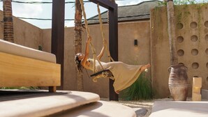 Une personne se balance sur une corde dans une cour, où se trouvent un banc en bois et un grand pot.