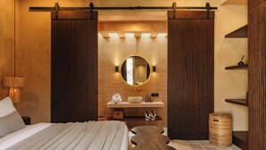 Une chambre à coucher comprenant un lit, un miroir rond, un banc en bois, une petite table et une étagère fixée au mur.