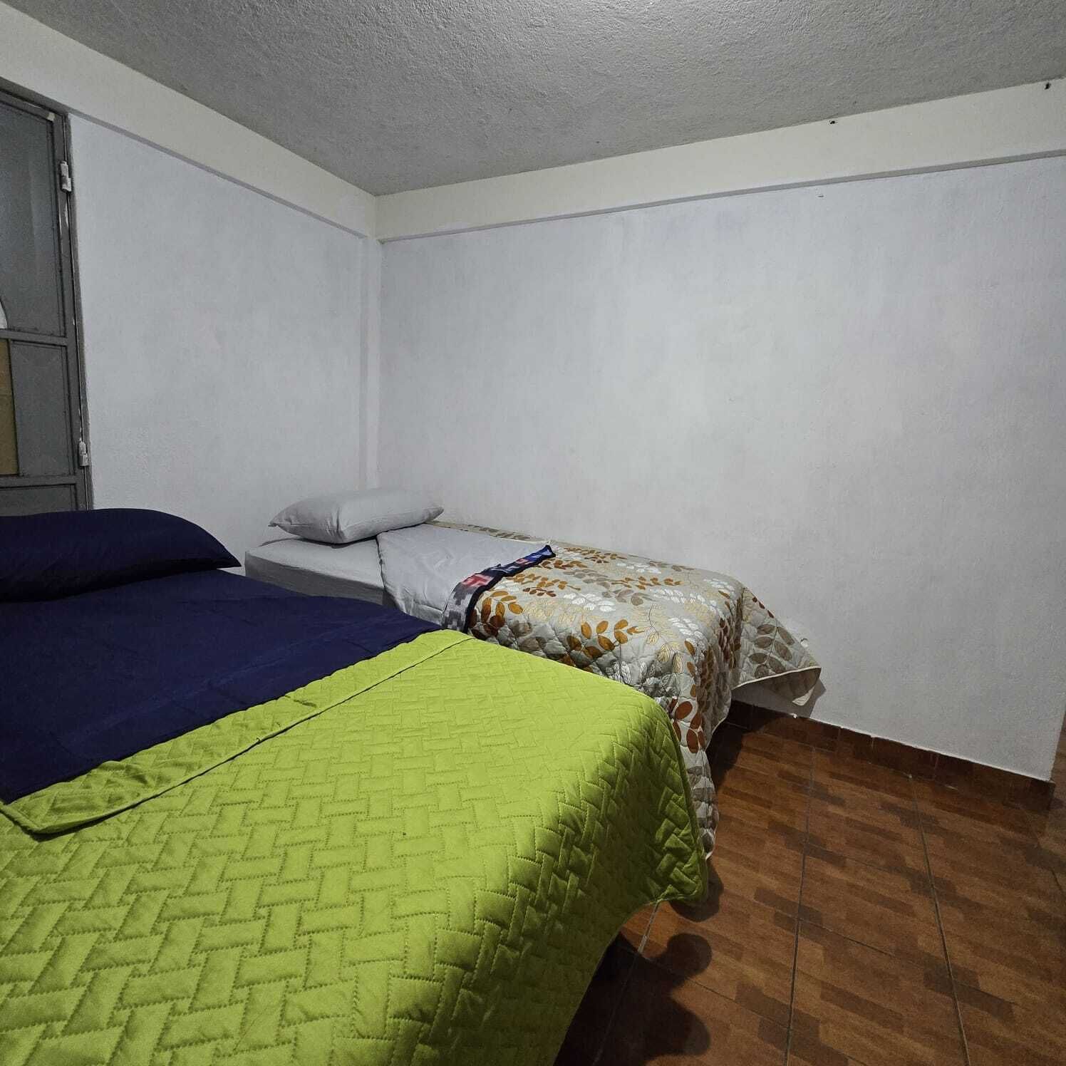 A small, sparsely furnished room with two beds, a window, and a wooden floor.