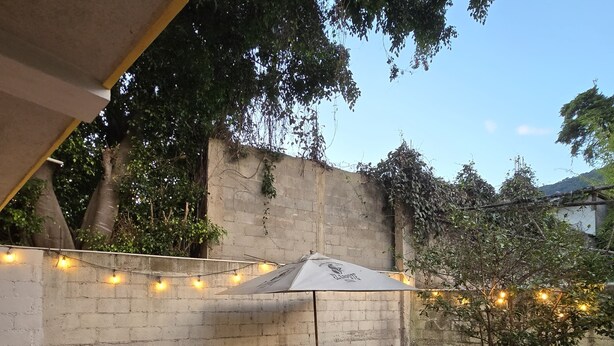 A patio area with a table and chairs under a white umbrella, surrounded by a low white wall and string lights. - Apartamentos el viajero (Panajachel)