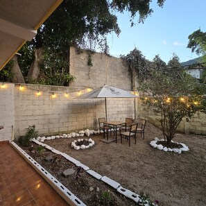 Ein Patio mit Tisch und Stühlen unter einem weißen Sonnenschirm, umgeben von einer niedrigen weißen Mauer und Lichterketten.