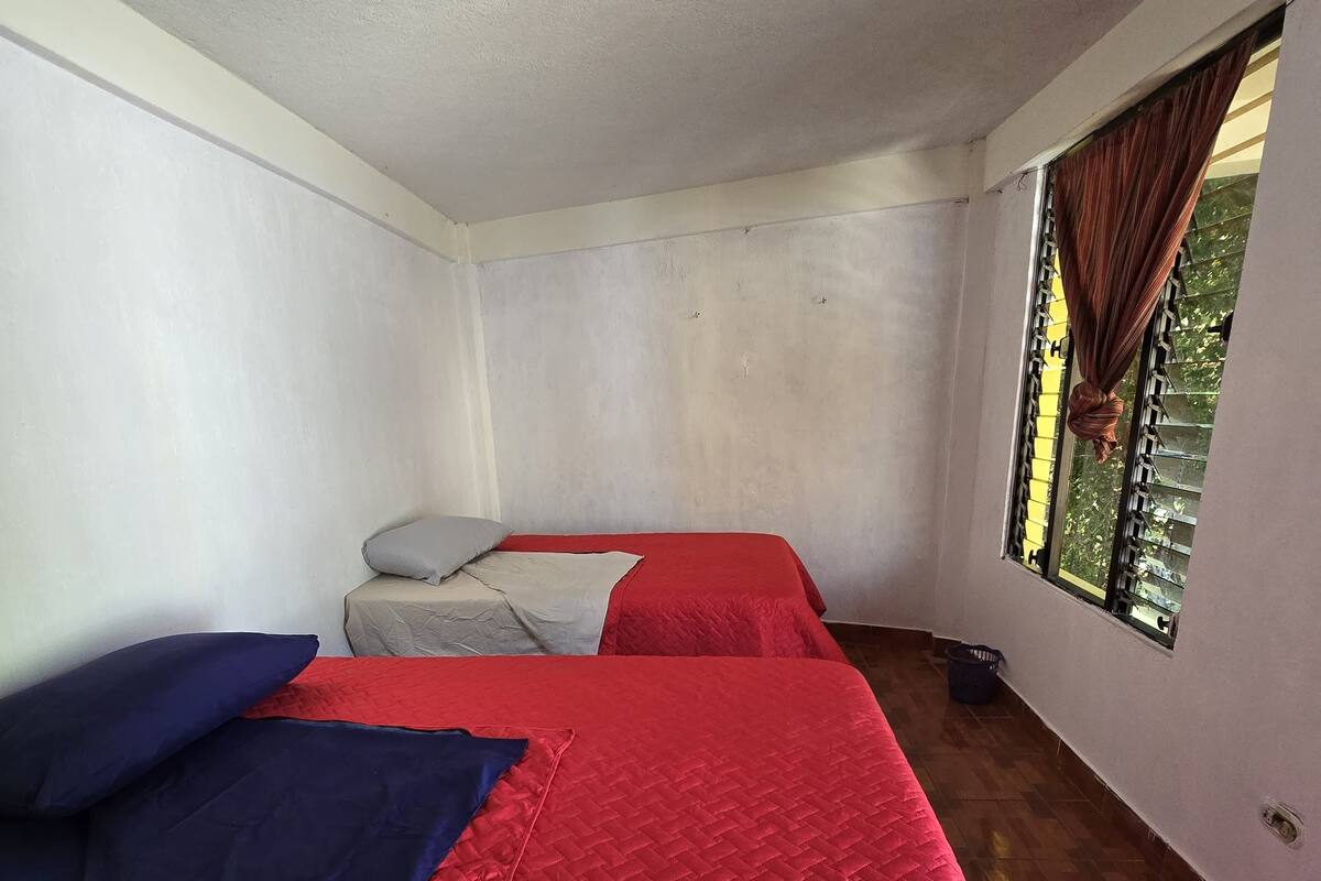 A room with two beds, each with a red quilted bedspread and pillows. The room has plain white walls and a window with curtains.