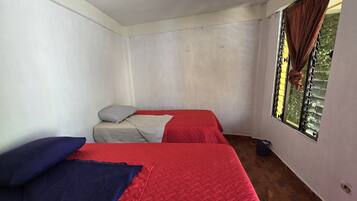 Ein Zimmer mit zwei Betten, jeweils mit einer roten gesteppten Bettdecke und Kissen. Die Wände sind schlicht weiß gestrichen und es gibt ein Fenster mit Vorhängen.
