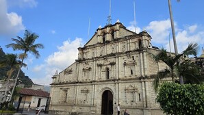 A historic stone building with a central doorway and a bell tower, surrounded by palm trees and a cobblestone plaza. - Apartamentos el viajero (Panajachel)