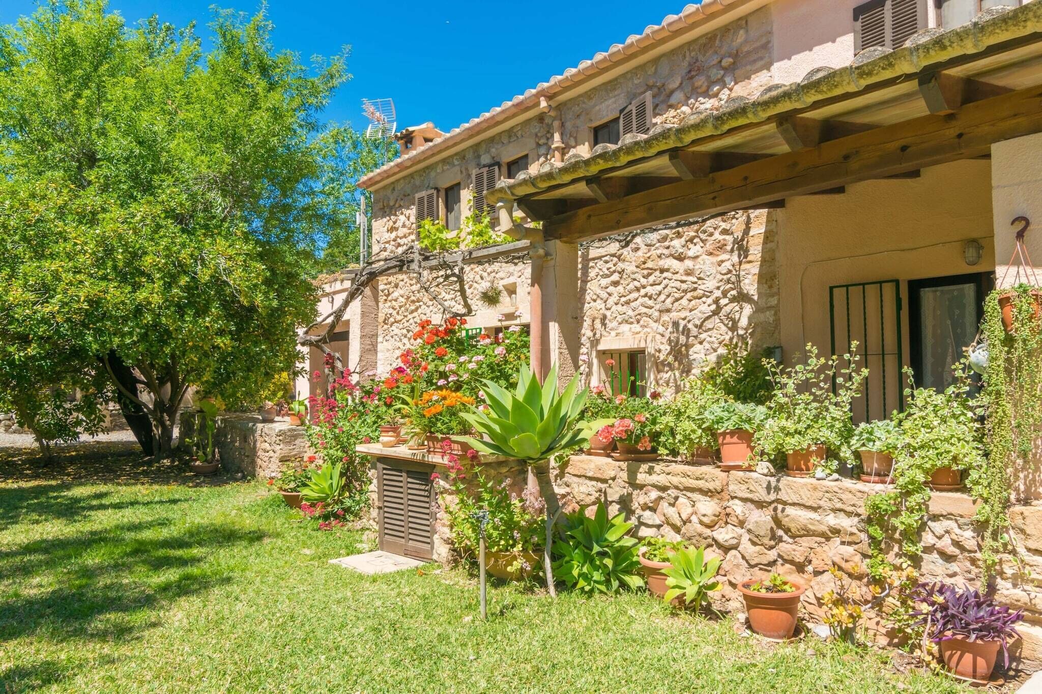 Edifici de pedra amb un porxo cobert, envoltat d'un jardí ben cuidat ple de plantes i flors en testos.