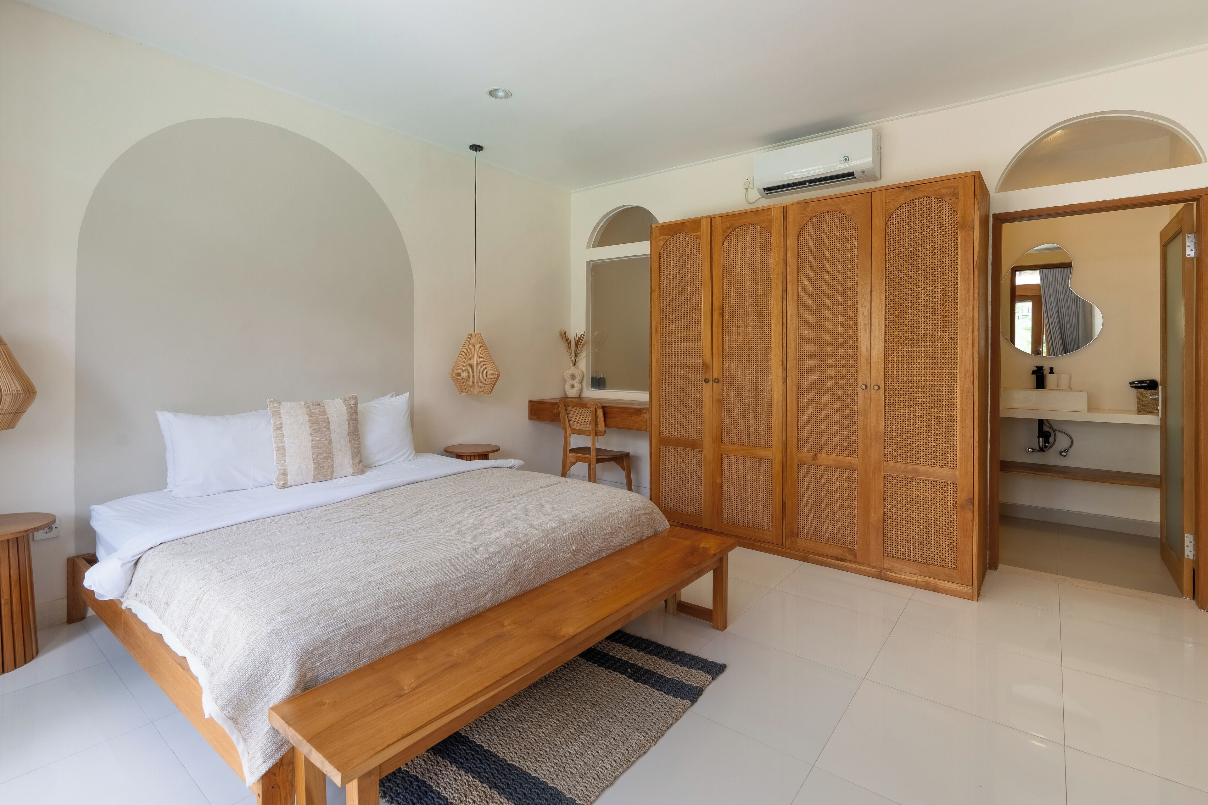 Un dormitorio con cama de madera, un banco, una lámpara colgante redonda, un armario y un baño con espejo y lavabo.