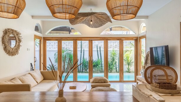 Ein geräumiger Wohnbereich mit einem Holztisch, einer Couch und einem Fernseher. Große Fenster bieten Blick auf einen Pool und Grünflächen.