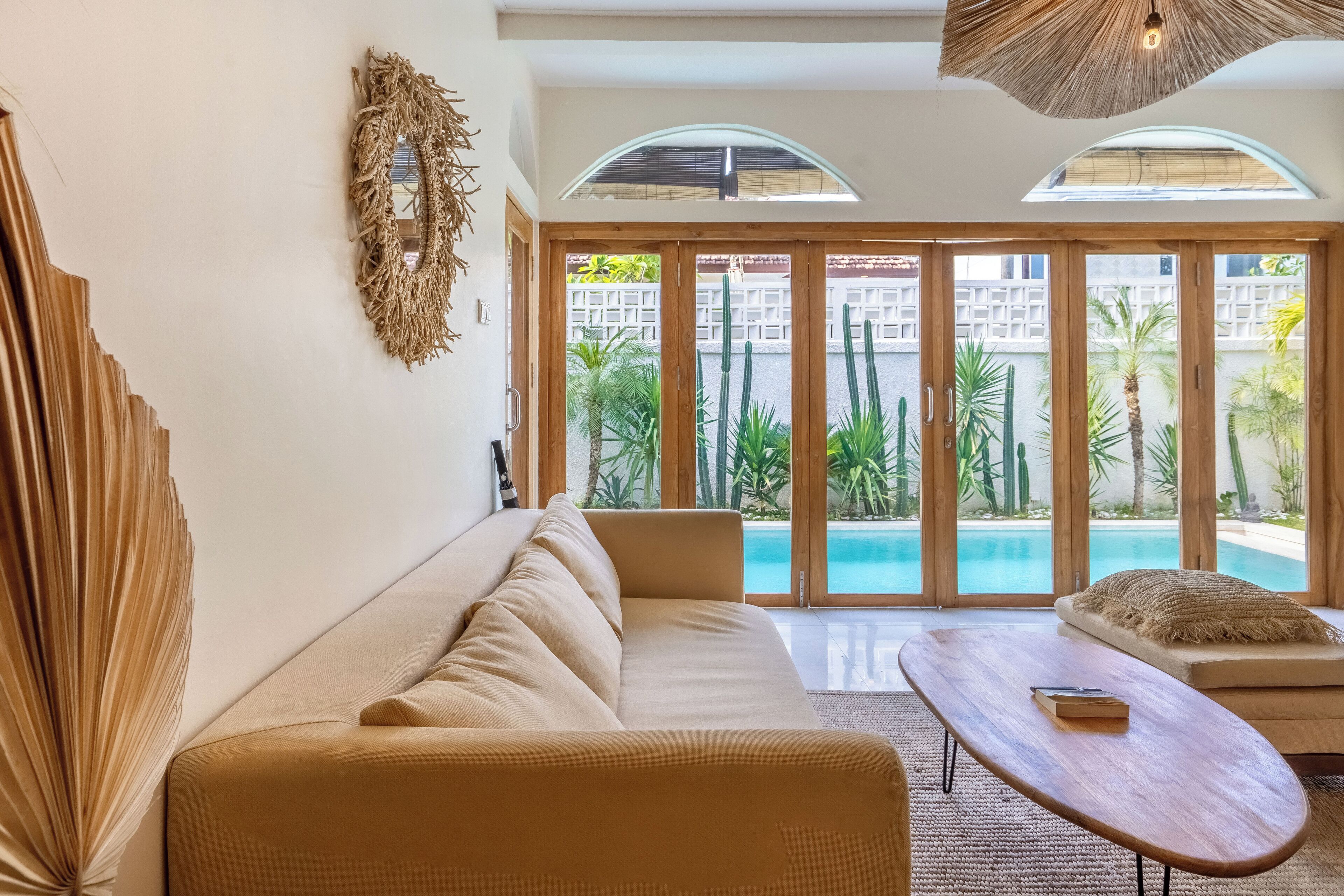 Ein modernes Wohnzimmer mit einem beigen Sofa, einem Holztisch und Blick auf einen Swimmingpool durch große Glasüren.