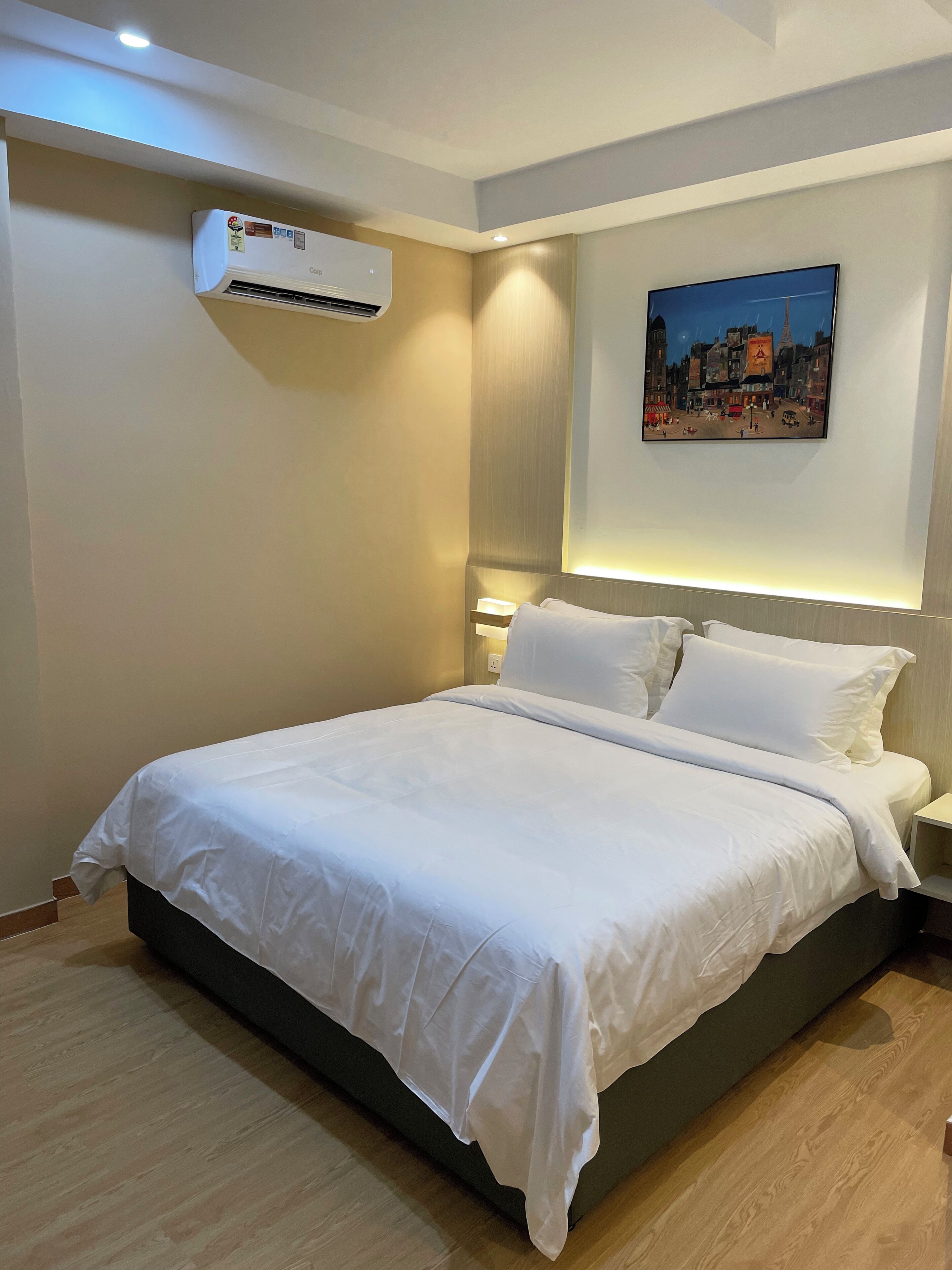 A neatly made bed with white linens, a wall-mounted air conditioner, a nightstand with a lamp, and a cityscape painting on the wall.