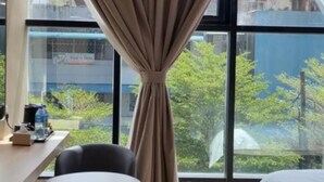 Een hotelkamer met een bed, een eettafel met stoelen en uitzicht op gebouwen en bomen door grote ramen.