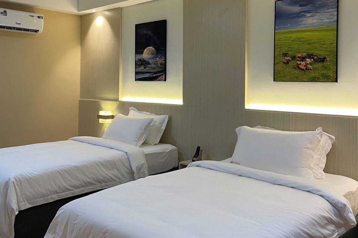 A hotel room with two single beds, a wall-mounted air conditioner, and two framed pictures.