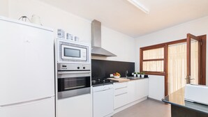 Una cucina moderna con mobili bianchi, forno, microonde e piano cottura integrati. Si nota una finestra con persiane in legno e una porta con pannello in vetro.