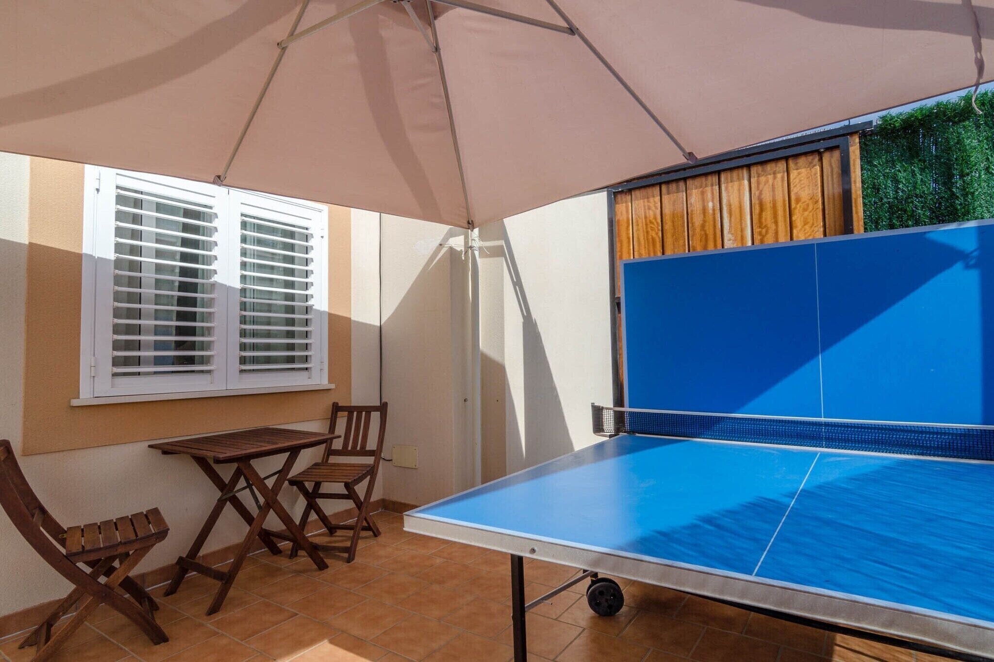A ping pong table under a canopy, a window with blinds, and a wooden table and chairs.