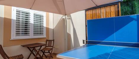 A ping pong table under a canopy, a window with blinds, and a wooden table and chairs.