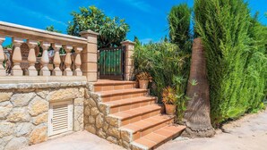 A stone staircase leading to a gated entrance, surrounded by lush greenery and a clear blue sky.