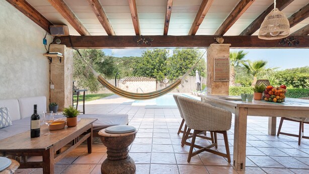 A covered outdoor patio with a wooden table, wicker chairs, a hammock, and a view of a pool and greenery. - Ca Na Rosa - Villa With Private Pool In Campanet (Campanet, Illes Balears)