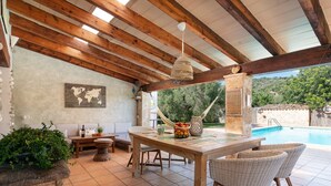 An outdoor patio with a wooden table, wicker chairs, a hanging light fixture, and a pool in the background. - Ca Na Rosa - Villa With Private Pool In Campanet (Campanet, Illes Balears)