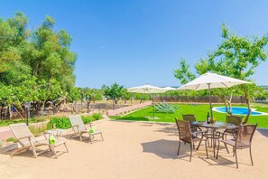 A patio area with lounge chairs, a table, and umbrellas, overlooking a swimming pool and landscaped garden. - Can Potdir - Villa With Private Pool In Ariany (Ariany, Illes Balears)