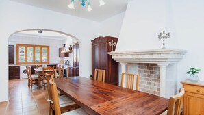 A spacious dining area with a long wooden table and chairs, a fireplace, and a kitchen visible through an archway.