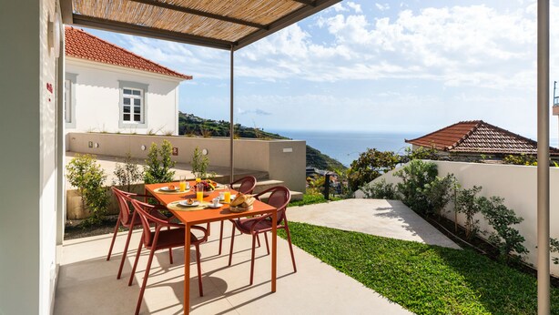 An outdoor dining area with a table and chairs, a view of the sea, and a building with a red roof. - Beautiful villa with rooftop pool | CarmHim (Calheta)