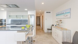 A modern kitchen with a central island, a dining area with a white table and chairs, and a hallway leading to another room. - Bon Estar - Apartment In Port D'alcúdia (Port D'alcúdia, Illes Balears)