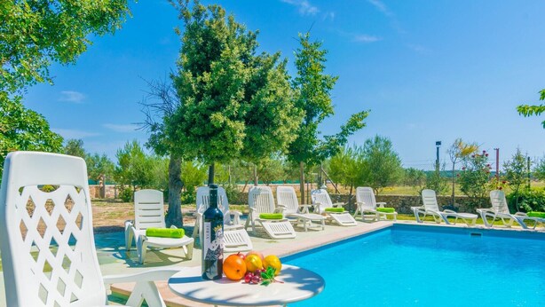 A poolside area with white lounge chairs, a table with fruits and a bottle, and a clear blue sky.