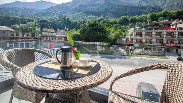 Un balcon avec des fauteuils et une table en osier, offrant une vue sur un paysage montagneux et sur un bâtiment d’hôtel.