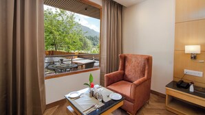 Une chambre d’hôtel avec une table à manger dressée pour deux personnes, un fauteuil confortable, une petite table de chevet avec une lampe, et une vue sur la verdure à l’extérieur.