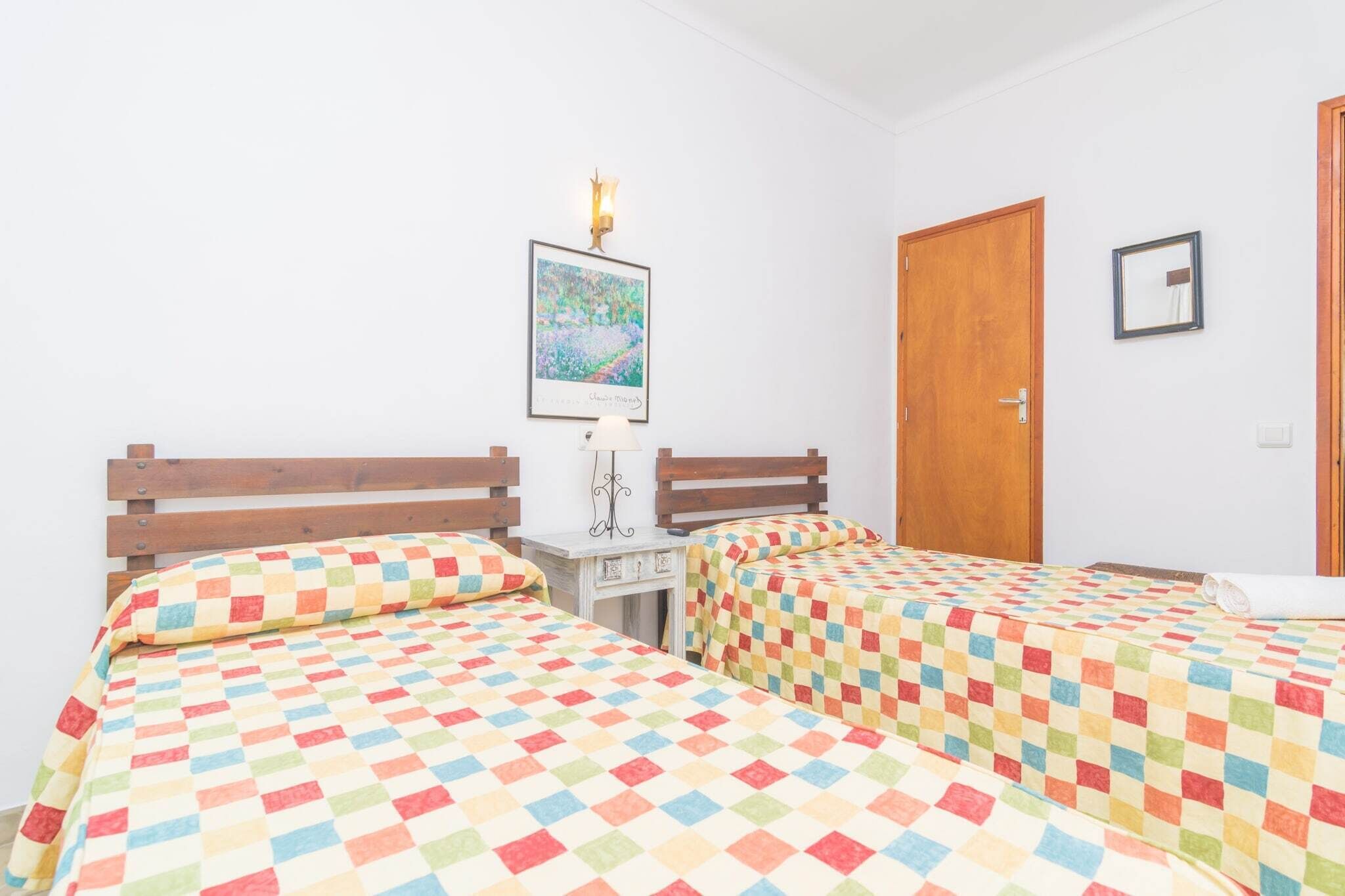 Habitació amb dos llits, cadascun amb un llençol de quadres de colors, una taula petita amb una làmpada, un quadre amb marc a la paret i una porta de fusta.