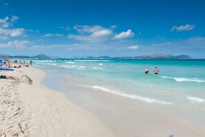 Ein Strand mit klarem türkisblauem Wasser, sandigem Ufer und Bergen in der Ferne.
