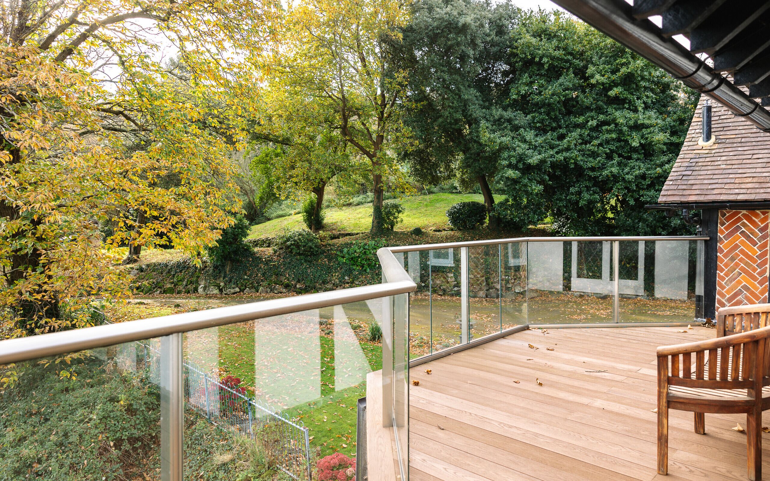 A wooden deck with a glass railing overlooking a landscaped garden with trees and a lawn.