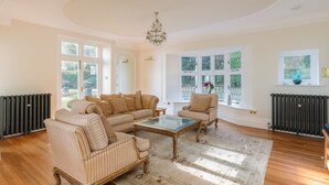 A spacious living room with a large window, a chandelier, and a radiator. - Luccombe Chine House (Shanklin)