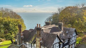 A traditional Tudor-style house with multiple chimneys, surrounded by lush greenery and overlooking a body of water. - Luccombe Chine House (Shanklin)