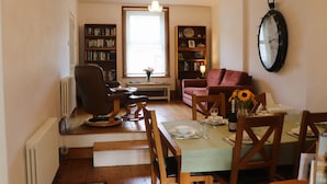 Un comedor con una mesa dispuesta para dos personas, una estantería, un sofá rojo y un reloj de pared.