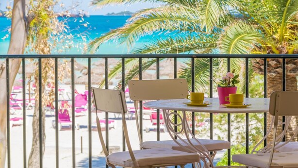 Een balkon met uitzicht op een strand en de oceaan. Er staan stoelen en een tafel met gele bekers en een roze bloempot.