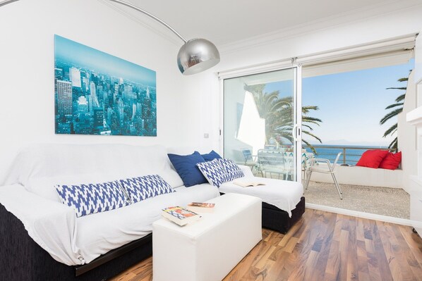 Un salon moderne avec un tableau représentant un paysage urbain, un canapé agrémenté de coussins à motifs, une table basse avec des livres, et un balcon offrant une vue sur la mer.